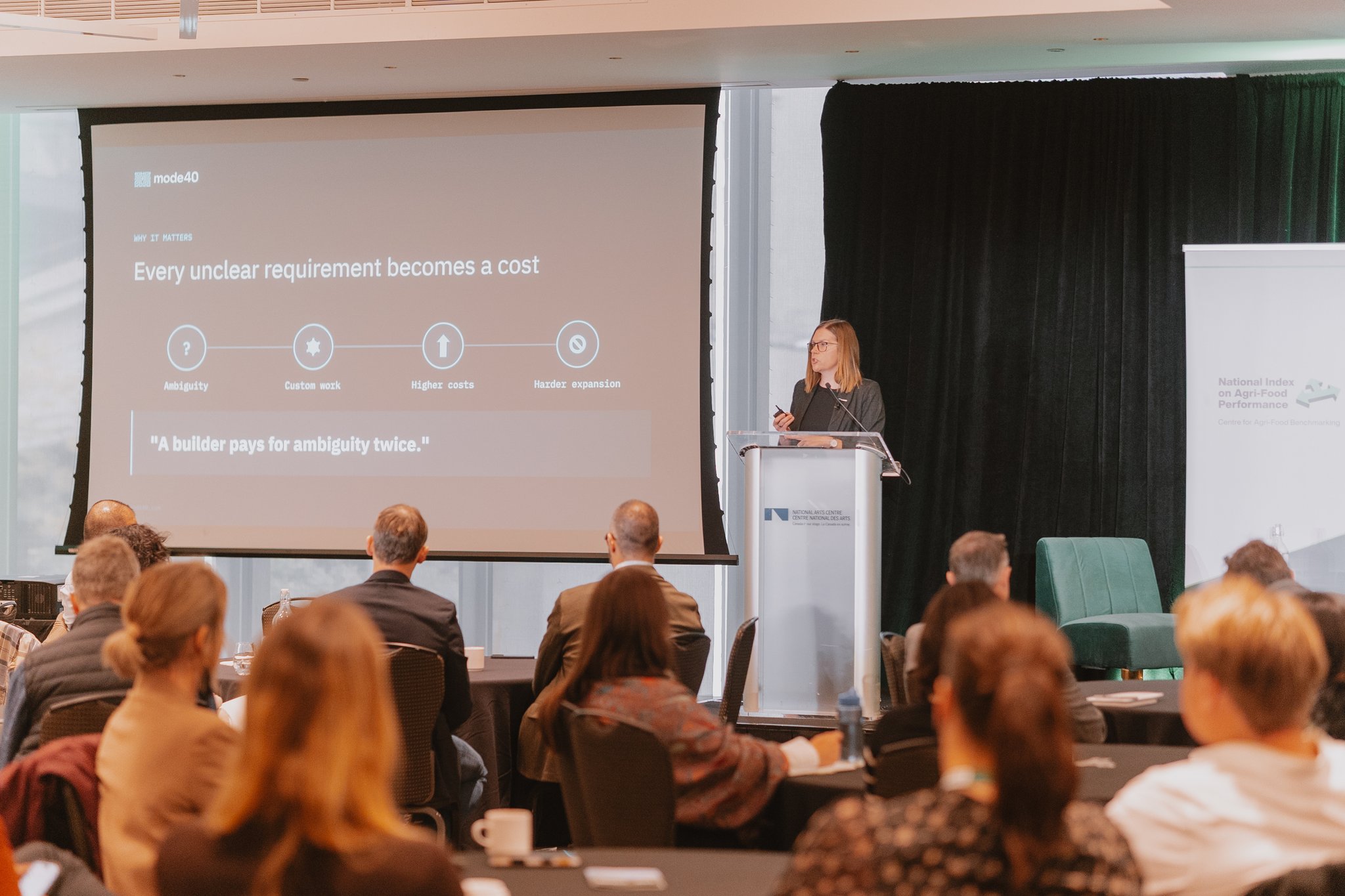 Tracy Gromniski at the podium beside a slide reading Every unclear requirement becomes a cost, with the audience in the foreground
